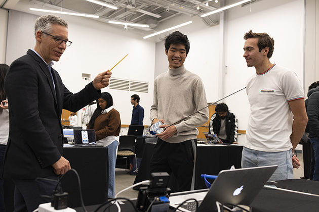 three men standing in a classroom holding a discussing fencing lances
